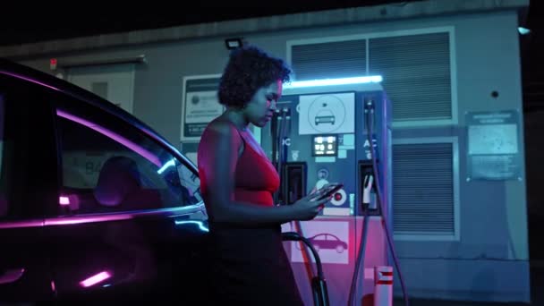 夜間のEvステーションでの電気自動車充電の横に立っている若いアフリカ系アメリカ人女性の中ショットを追跡します 彼女は携帯電話を使って待ってる — ストック動画
