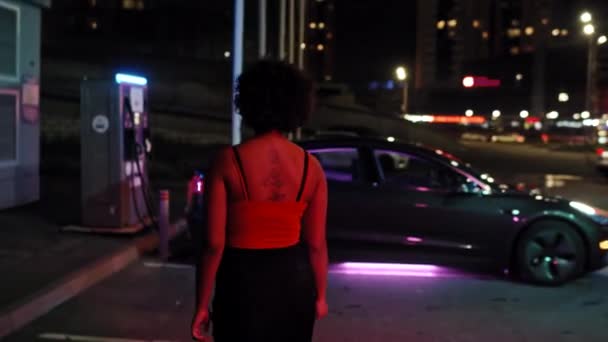 電気自動車を充電するために夜にEvステーションに向かって歩く若いアフリカ系アメリカ人女性の背面ビューで撮影 — ストック動画