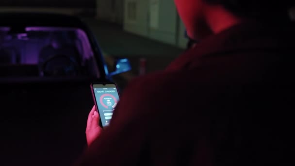 アフリカ系アメリカ人の若い女性の肩のショットを追跡する彼女の携帯電話でアプリを見て彼女の電気自動車は夜にEvステーションで充電しながら — ストック動画