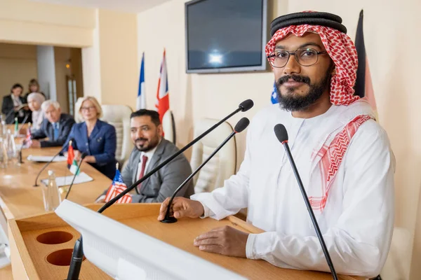 Jeune orateur arabe regardant la caméra tout en étant debout par tribune — Photo