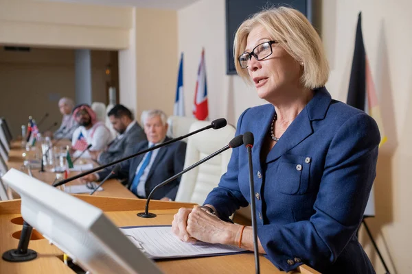 Madura rubia delegada en ropa formal hablando en micrófono — Foto de Stock