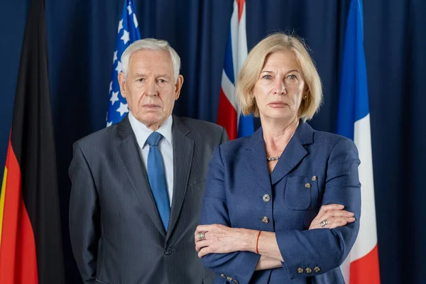 Two mature political leaders in formalwear standing against four flags — Stockfoto