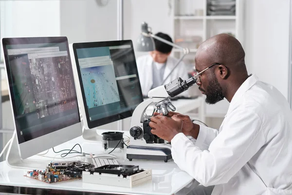 Joven hombre africano serio con microscopio sentado por el lugar de trabajo — Foto de Stock