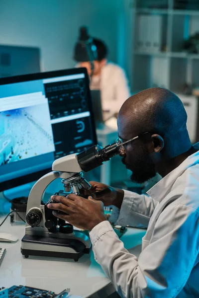 Joven de etnia africana sentado en el lugar de trabajo y mirando al microscopio — Foto de Stock