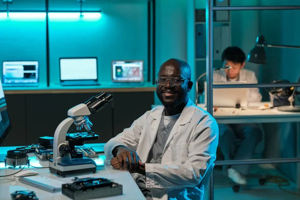 Feliz joven científico africano con microscopio sentado en el lugar de trabajo — Foto de Stock