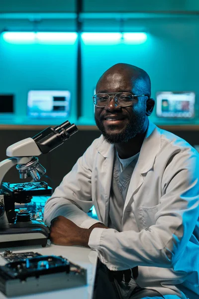 Feliz joven trabajador de laboratorio sentado junto al escritorio con microscopio — Foto de Stock