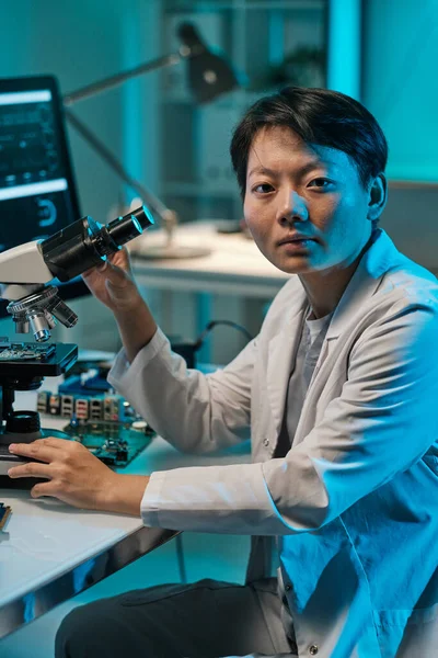 Young Chinese woman in whitecoat looking at camera while working with microscope — Stock Photo, Image