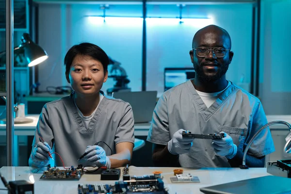 Twee jonge succesvolle technici in handschoenen en uniform werken per bureau — Stockfoto