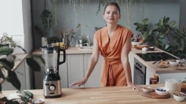 Sağlıklı yaşam tarzına sahip genç bir kadının portresi sıcak mutfağa blender ve meyve püresi için hazırlanmış malzemelerle giriyor. Mutfak tezgahına yaslanıyor ve kameraya gülümsüyor.