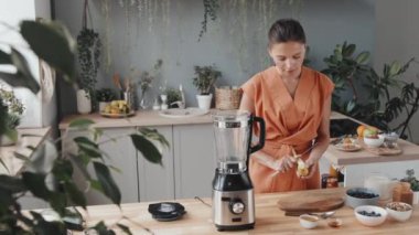 Sağlıklı yaşam tarzı olan genç bir kadının muz soyarken ve kesmek için tahta tahtaya yerleştirirken görüntüsü. Mutfak tezgahında taze meyve, blender, fındık ve sebze var.