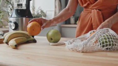 Mutfaktaki poşetten taze meyve çıkaran tanınmayan genç bir kadının el bilgisayarı. Blender mutfak tezgahında duruyor.