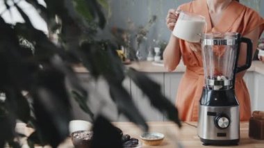 Sıcak mutfakta taze meyveyle smoothie yaparken, blender 'a süt döken genç bir kadının orta bölüm takibi.