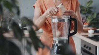 Genç bir kadının, mutfakta sütle meyve püresi yaparken muz keserken orta bölüm izleme görüntüsü.