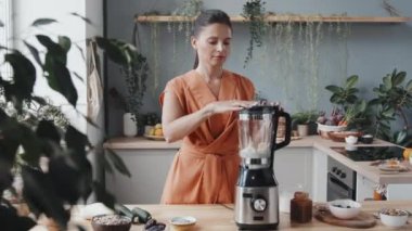 Genç vejetaryen bir kadının bol güneş ışığı ve yeşil bitkilerle sıcacık mutfakta sağlıklı meyve püresi yaparken görüntüsü.