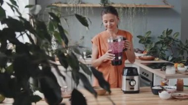 Genç vejetaryen bir kadının sıcak mutfakta bol güneş ışığı ve yeşil bitkilerle blenderdan lezzetli içecekler dökerkenki görüntüsü.