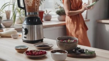 Mutfakta pencerenin önünde duran ve cep telefonu kullanan tanınmayan genç bir kadının orta bölümünün izini sürmek. Lezzetli smoothie kasesi, blender ve taze malzemeler mutfak tezgahında.
