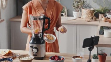 Tanınmayan genç bir kadının orta bölüm takibi taze meyveyi karıştırıcıya koyuyor ve rahat bir mutfakta bitkilerle kendini cep telefonuyla filme çekiyor.