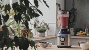 Sıcak mutfakta bol güneş ışığı ve bitkilerle birlikte mikserde dönen pembe smoothie 'nin orta boy görüntüsü. Taze meyve kaselerde tezgahta.