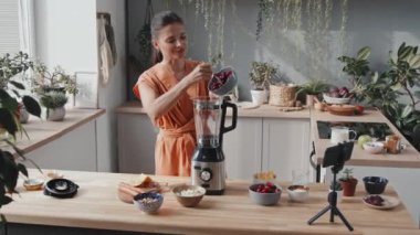 Yemek blogu için video çeken neşeli genç bir kadının izini sürüyorum. Konuşuyor, çilek, kiraz ve portakal dilimlerini blender 'a koyuyor ve sıcak mutfakta bitkilerle smoothie yapıyor.