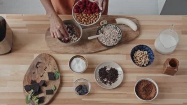 Lezzetli meyve suyuna ahududu ekleyen tanınmayan genç bir kadının el kamerasıyla çekilmiş fotoğrafı. Taze meyveler, kakao, koyu çikolata, fındık, incir, süt ve hindistan cevizi taneleri masada.