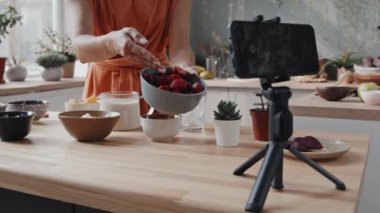 Kendini cep telefonuyla kaydeden ve ev yapımı smoothie için çeşitli malzemeler gösteren tanınmayan genç bir kadının orta bölüm çekimi.