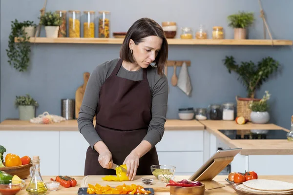 Young female in apron watching video recipe while cutting capsicum — Foto Stock