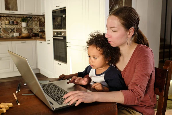 Fiatal elfoglalt nő fogja az aranyos kis fia, miközben ül a laptop előtt — Stock Fotó