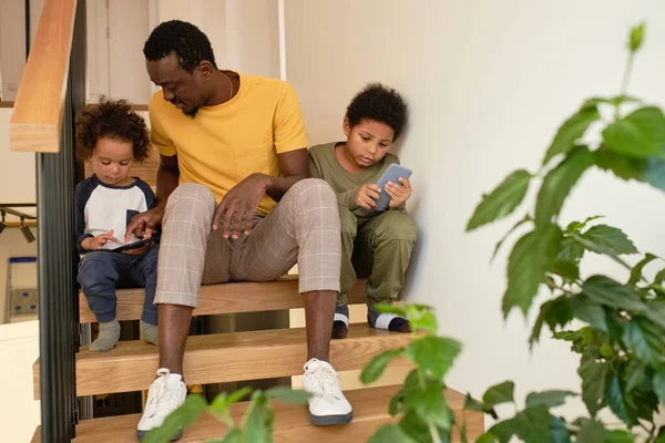 Ung man av afrikansk etnicitet pratar med en av sina söta söner med smartphones — Stockfoto