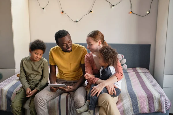 Familia joven feliz de cuatro sentados en la cama doble, viendo vídeo en línea y discutiéndolo —  Fotos de Stock