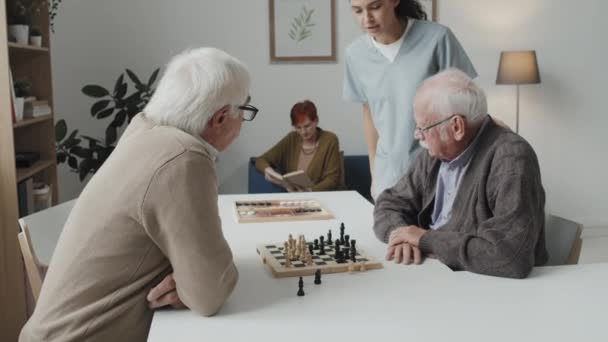 介護中の若い女性看護師が老人ホームに住む高齢者と話をする様子 男遊びチェスながら女性読み本でソファ — ストック動画