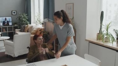 Orta boy kızıl saçlı yaşlı bir kadın huzurevinde oturmuş, örgü örerken genç bir hemşire ona tek kullanımlık bardakta haplar getiriyor ve sağlığını soruyor.
