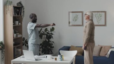 Orta boy beyaz bir adam, halterli fiziksel egzersiz yapıyor. Afro-Amerikan erkek hemşirenin önünde durup ona yardım ediyor.