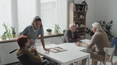 Genç hemşirenin huzur evindeki masada oturan yaşlı erkek ve kadına tek kullanımlık bardaklarda haplar verdiği orta boy bir fotoğraf.