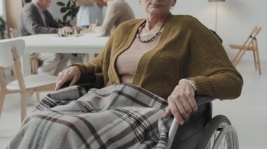 Tekerlekli sandalyede oturan ve huzurevindeki kameraya bakan yaşlı kızıl saçlı kadının portresini kaldır.