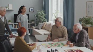Orta sınıf kadın hemşire, beyaz yaşlı adamı huzurevinde oturmuş masa başında masa başında masa oyunları oynayan yaşlı insanlarla tanıştırıyor.