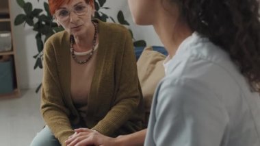 Yetişkin kızıl saçlı kadın ve destekleyici genç hemşirenin el ele tutuştuğu, huzurevindeki kanepede oturduğu bir fotoğraf.