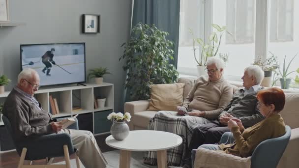中低速ショットの4人の高齢者が一緒に余暇を過ごすテレビで居心地の良い老人ホームのリビングルーム 読書本 編み物や会話を持っています — ストック動画
