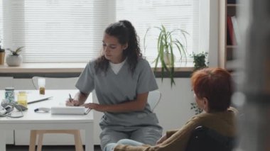 Orta boylu, kızıl saçlı, tekerlekli sandalyedeki yaşlı bir kadının modern ofisteki randevusu sırasında genç bir bayan doktorla konuşması.