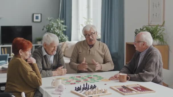 中出し4現代白人高齢者のテーブル遊びボードゲームに座っていると居心地の良いリビングルームで会話を持っている — ストック動画