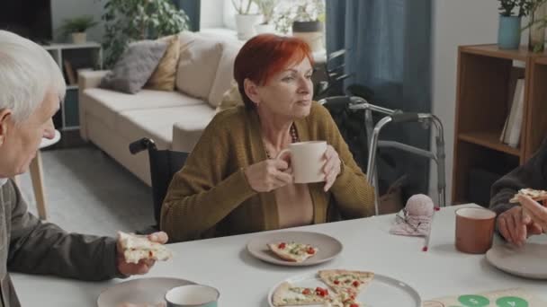 車椅子でピザを食べ テーブルで他の高齢者と話をし 老人ホームで昼食を共にした赤髪の白人のシニア女性の中ショット — ストック動画