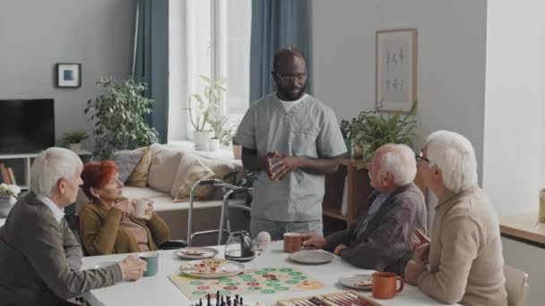 昼食時に高齢者のグループが一緒に座って会話をしているテーブルのそばに立つアフリカ系アメリカ人男性看護師の中程度のショット — ストック動画