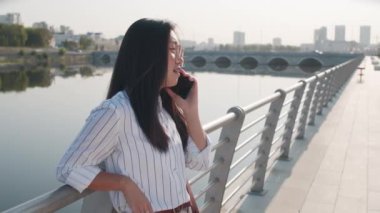 Güneşli bir günde nehir kenarında duran akıllı telefondan konuşan genç ve güzel bir Asyalı kadının orta boy fotoğrafı.