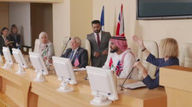 Orta yaşlı erkek politikacının basın toplantısına katıldığı, farklı ülkelerin diğer siyasi liderleriyle masaya oturduğu ve mikrofonla dinleyicileri selamladığı orta boy bir fotoğraf.