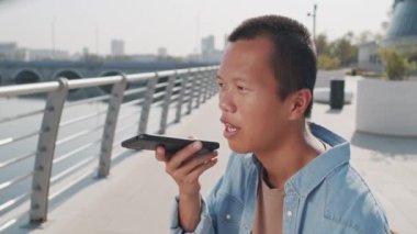 Genç Asyalı bir adamın cep telefonuyla sesli mesaj kaydettiği orta ölçekli bir fotoğraf. Yaz günü nehir kenarında oturuyor.