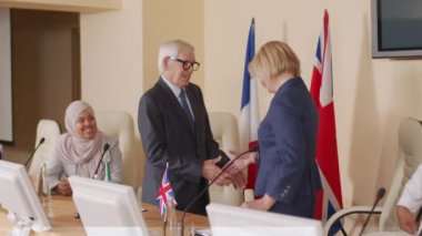 İki gülümseyen beyaz erkek ve kadın politikacının büyük salonda düzenlenen basın toplantısında diğer siyasi liderler ve seyircilerin önünde tokalaşırken orta ölçekli bir fotoğraf.