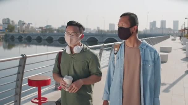 晴れた日に川の堤防に沿って歩く犠牲者と顔のマスクで2人の若いアジアの男性の中のショットを追跡会話を持って — ストック動画