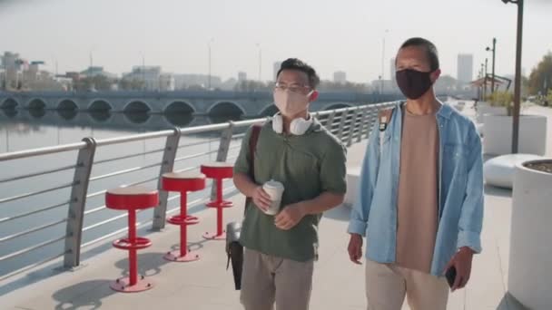 暖かい晴れた日に川の堤防に沿って歩く顔のマスクで2人の若いアジアの男性の友人の中のショットを追跡 — ストック動画