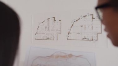 Duvarda asılı ev planına bakan iki genç mimarın zoom-out görüntüsü. Ellerindeki çizimle karşılaştırıyor.