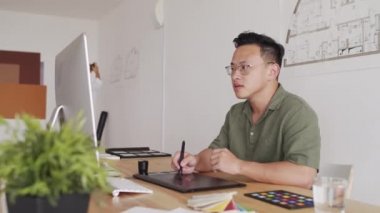 Genç Asyalı erkek mimarın grafik tablete çizim yaptığı orta boy bir fotoğraf. Sonra kameraya gülümseyip modern ofisteki bilgisayarın önünde oturuyor.