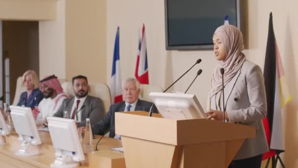 ヒジャーブ州の若いイスラム教徒の女性政治家が 大きな会議室に立っている間に聴衆に話しているのを撮影しました 彼女のスピーチを聞くテーブルに座っている他の国の代表 — ストック動画